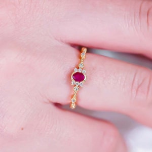 Może przedstawiać: Złoty pierścionek z owalnym rubinem pośrodku. Pierścionek ma delikatny wzór z małymi, przezroczystymi kamieniami akcentującymi i skomplikowanymi detalami wzdłuż obrączki. Pierścionek jest pokazany na palcu.