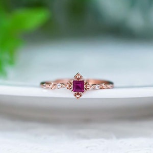 May include: A delicate rose gold ring featuring a square ruby gemstone surrounded by smaller clear gemstones. The ring has a vintage-inspired design with intricate details.