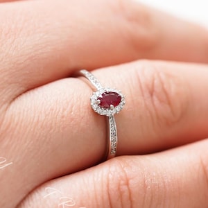 Pode incluir: Um delicado anel de prata com uma gema oval vermelha cercada por diamantes menores. O anel está em um dedo.