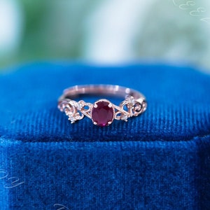 Puede incluir: Un anillo de oro rosa con una piedra preciosa de rubí y pequeños diamantes engastados en un delicado diseño floral. El anillo se muestra en una caja de anillo de terciopelo azul.
