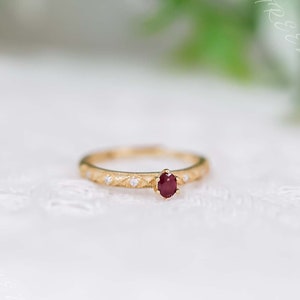 May include: A delicate gold ring with a small, oval-shaped ruby gemstone set in the center. The band is adorned with tiny, sparkling diamonds.