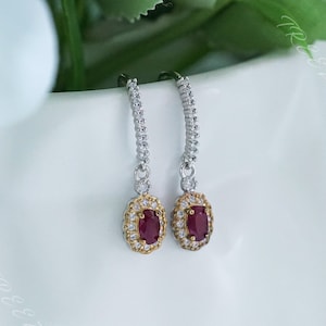 Könnte beinhalten: Ein Paar silberne Ohrhänger mit einem roten ovalen Edelstein, der von einem Heiligenschein aus kleineren weißen Edelsteinen umgeben ist. Die Ohrringe haben einen goldenen Akzent um den Edelstein.