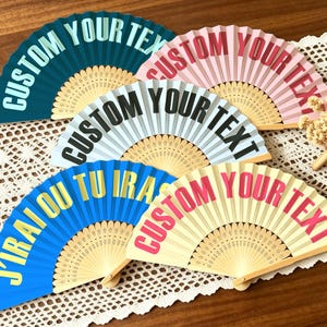 May include: Five folding hand fans with wooden ribs and fabric leaves in teal, pink, light blue, bright blue, and cream. The fans feature the text CUSTOM YOUR TEXT in various colours, while the bright blue fan displays the text J'IRAI OU TU IRAS.
