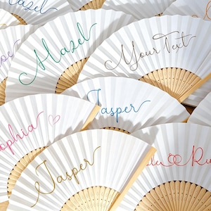 Op de afbeelding: Een verzameling witte vouwwaiers met lichtgekleurde houten ribben. Elke waaier is voorzien van gepersonaliseerde namen of tekst in verschillende gekleurde cursieve lettertypes, waaronder Hazel, Jasper, Sophia en Ruby, met soms kleine hart- of ringpictogrammen.