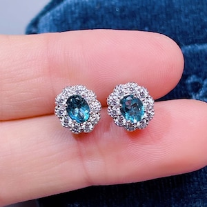 May include: A pair of silver stud earrings with a blue oval gemstone surrounded by a halo of smaller white gemstones.