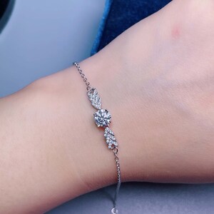 May include: A delicate silver bracelet featuring a central round, clear gemstone, flanked by two decorative leaf-shaped elements, each adorned with smaller, sparkling stones. The bracelet is designed with a fine chain.
