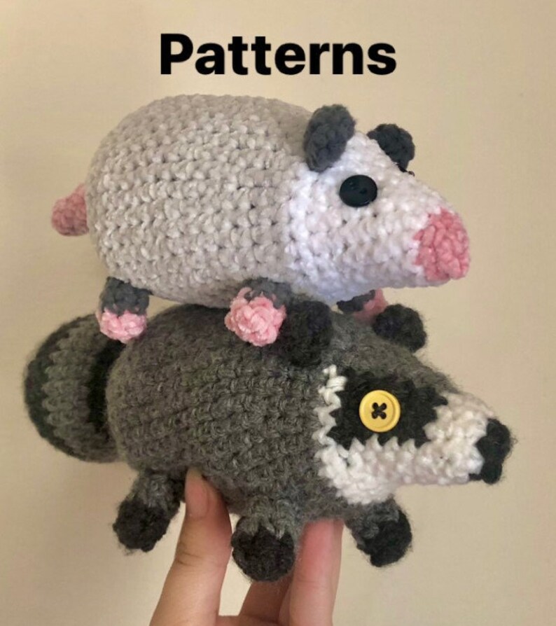 May include: A crocheted gray raccoon plush toy with a white face and a yellow button for an eye. A crocheted gray and white opossum plush toy with pink accents is sitting on top of the raccoon.