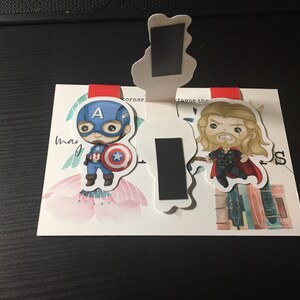 Op de afbeelding: Twee magnetische bladwijzers met cartoonillustraties van Captain America en Thor. Captain America draagt zijn kenmerkende blauwe en rode pak en houdt zijn schild vast. Thor draagt zijn rode cape en houdt zijn hamer vast. De bladwijzers bevinden zich op een witte achtergrond met een bloemmotief.