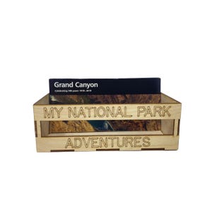 May include: A wooden crate with the text "My National Park Adventures" engraved on the front. A postcard with the text "Grand Canyon Celebrating 100 years: 1919-2019" is inside the crate.