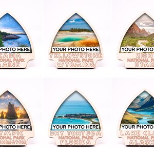 May include: Six wooden arrow-shaped signs with a photo cutout in the center. Each sign features a different national park in the United States, including Acadia National Park in Maine, Yellowstone National Park in Wyoming, Zion National Park in Utah, Olympic National Park in Washington, Dry Tortuga National Park in Florida, and Lake Clark National Park in Alaska. The signs are blank where the photos should be, and the text "Your Photo Here" is printed below each photo cutout.