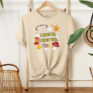 Op de afbeelding: Een beige t-shirt met korte mouwen en een grafische print met de tekst "I KNEW IT, I KNEW YOU", een kralenarmband, een gele sheriffster, een rode cowboylaars, een rode cowboyhoed, een gele ster en de letters "TS5" in gekleurde blokken.