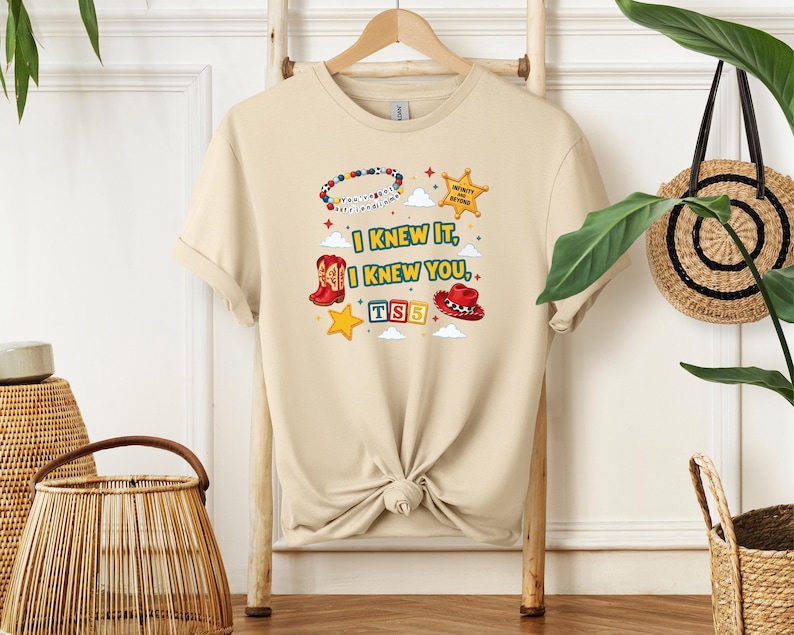 Op de afbeelding: Een beige t-shirt met korte mouwen en een grafische print met de tekst "I KNEW IT, I KNEW YOU", een kralenarmband, een gele sheriffster, een rode cowboylaars, een rode cowboyhoed, een gele ster en de letters "TS5" in gekleurde blokken.