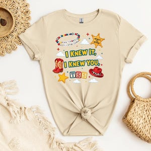 Op de afbeelding: Een beige t-shirt met korte mouwen en een grafische print met de tekst I KNEW IT, I KNEW YOU, TS5 en INFINITY AND BEYOND. Het ontwerp bevat een kralenarmband, een sheriffster, rode cowboylaarzen, een rode hoed en blokken met letters.