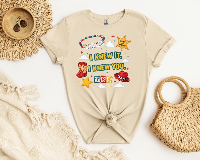 Op de afbeelding: Een beige t-shirt met korte mouwen en een grafische print met de tekst I KNEW IT, I KNEW YOU, TS5 en INFINITY AND BEYOND. Het ontwerp bevat een kralenarmband, een sheriffster, rode cowboylaarzen, een rode hoed en blokken met letters.