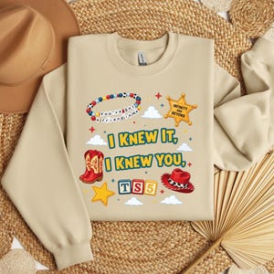 Op de afbeelding: Een beige sweater met ronde hals en een grafische print met de tekst I KNEW IT, I KNEW YOU, TS5, INFINITY AND BEYOND en YOU'VE GOT A FRIEND IN ME. Het ontwerp bevat een kralenarmband, een sheriffster, cowboylaarzen en een cowboyhoed.