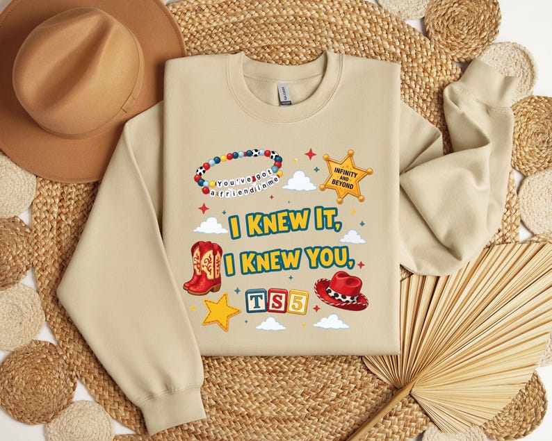 Op de afbeelding: Een beige sweater met ronde hals en een grafische print met de tekst I KNEW IT, I KNEW YOU, TS5, INFINITY AND BEYOND en YOU'VE GOT A FRIEND IN ME. Het ontwerp bevat een kralenarmband, een sheriffster, cowboylaarzen en een cowboyhoed.