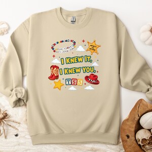 Op de afbeelding: Een beige sweater met ronde hals en een grafische print met de tekst "I KNEW IT, I KNEW YOU", "TS5" en "INFINITY AND BEYOND". Het ontwerp bevat een kralenarmband, een gele sheriffster, rode cowboylaarzen, een rode hoed en alfabetblokken.