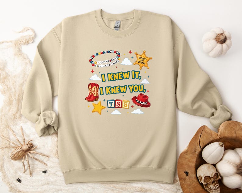 Op de afbeelding: Een beige sweater met ronde hals en een grafische print met de tekst "I KNEW IT, I KNEW YOU", "TS5" en "INFINITY AND BEYOND". Het ontwerp bevat een kralenarmband, een gele sheriffster, rode cowboylaarzen, een rode hoed en alfabetblokken.