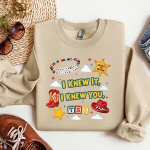 Op de afbeelding: Een beige sweater met ronde hals en een grafische print met de tekst I KNEW IT, I KNEW YOU, TS5, INFINITY AND BEYOND en YOU'VE GOT A FRIEND IN ME. Het ontwerp bevat een kralenarmband, een sheriffster, cowboylaarzen en een cowboyhoed.