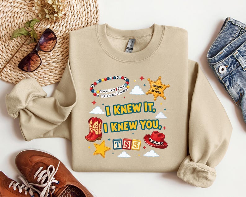 Op de afbeelding: Een beige sweater met ronde hals en een grafische print met de tekst I KNEW IT, I KNEW YOU, TS5, INFINITY AND BEYOND en YOU'VE GOT A FRIEND IN ME. Het ontwerp bevat een kralenarmband, een sheriffster, cowboylaarzen en een cowboyhoed.