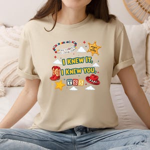 Op de afbeelding: Een beige t-shirt met een grafische print met de tekst I KNEW IT, I KNEW YOU, TS5 en INFINITY AND BEYOND. Het ontwerp bevat een kralenarmband, een sheriffster, een rode cowboylaars, een rode cowboyhoed, gele sterren en witte wolken.