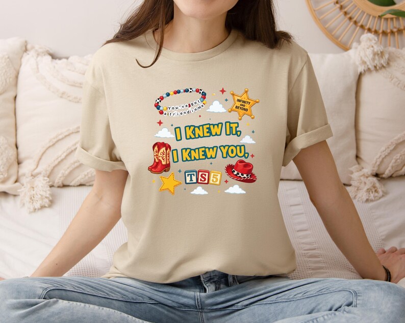 Op de afbeelding: Een beige t-shirt met een grafische print met de tekst I KNEW IT, I KNEW YOU, TS5 en INFINITY AND BEYOND. Het ontwerp bevat een kralenarmband, een sheriffster, een rode cowboylaars, een rode cowboyhoed, gele sterren en witte wolken.