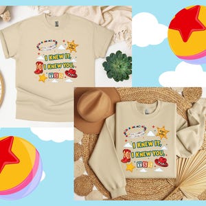 May include: A beige short-sleeve t-shirt and a beige long-sleeve sweatshirt featuring a graphic print with the text I KNEW IT, I KNEW YOU, TS5, and INFINITY AND BEYOND, alongside illustrations of a beaded bracelet, cowboy boots, a hat, and stars.