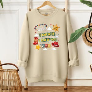 Op de afbeelding: Een beige sweater met ronde hals en een grafische print met de tekst "I KNEW IT, I KNEW YOU", een kralenarmband, een sheriffster, een rode cowboylaars, een rode cowboyhoed en "TS5" op kleurrijke blokken, omringd door kleine wolken.
