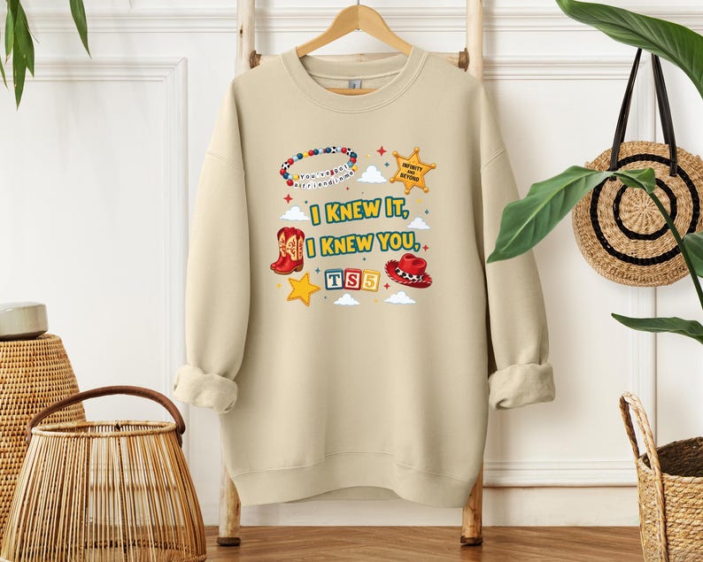 Op de afbeelding: Een beige sweater met ronde hals en een grafische print met de tekst "I KNEW IT, I KNEW YOU", een kralenarmband, een sheriffster, een rode cowboylaars, een rode cowboyhoed en "TS5" op kleurrijke blokken, omringd door kleine wolken.