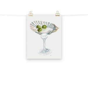 May include: A watercolor painting of a martini glass with three green olives in a seashell. The glass is clear and the seashell is white with a light pink and blue pattern.