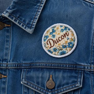 Peut inclure: Un écusson circulaire en tissu orné d'un motif floral de petites fleurs bleues et jaunes sur fond crème, avec le mot Ducon brodé en écriture cursive marron, fixé sur une veste en jean bleu avec des boutons métalliques et des coutures visibles.