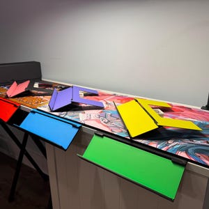 May include: Five colourful laptop stands in different colours, including red, blue, purple, yellow, and green, are arranged on a desk with a colourful anime-style design. The stands are made of a sturdy material and are designed to elevate laptops for a more ergonomic viewing experience.