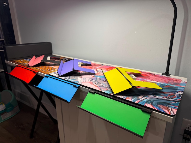 May include: Five colourful laptop stands in different colours, including red, blue, purple, yellow, and green, are arranged on a desk with a colourful anime-style design. The stands are made of a sturdy material and are designed to elevate laptops for a more ergonomic viewing experience.