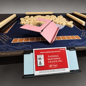 Puede incluir: Un juego de Mah Jongg sobre una mesa de madera oscura. El juego incluye fichas beige, una superficie de juego azul con dise&ntilde;os florales y un soporte rosa para fichas. Un letrero con el texto "NATIONAL MAH JONGG LEAGUE, INC." es visible.