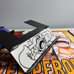 May include: A black and white drawing of a character with a large mouth and a cloud behind them. The drawing is on a white background and is inside a black frame. The frame is sitting on top of a colorful comic book page with the word "emperors" printed in yellow.