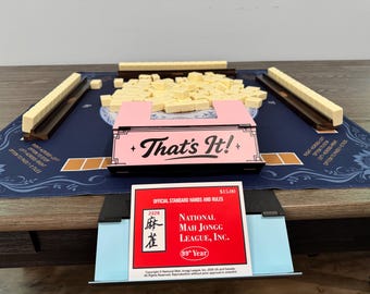 May include: A Mah Jongg set with cream-colored tiles, a pink tile holder with "That's It!" text, and a blue playing mat. The set includes racks and a rule card from the National Mah Jongg League, Inc.