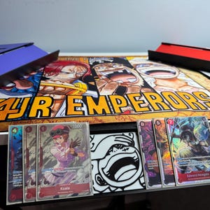 May include: Four Emperor playing cards featuring characters from the anime One Piece. The cards are laid out on a playmat with a colorful illustration of the four emperors. The cards feature the characters Koala and Edward Newgate.