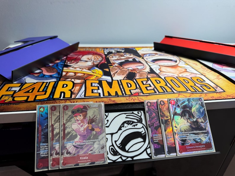 May include: Four Emperor playing cards featuring characters from the anime One Piece. The cards are laid out on a playmat with a colorful illustration of the four emperors. The cards feature the characters Koala and Edward Newgate.