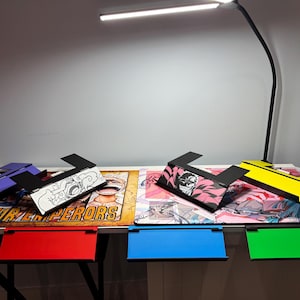 May include: Five different colored laptop stands with various anime designs. The stands are made of cardboard and fold flat for easy storage. The stands are on a table with a white surface. The text "FOUR EMPERORS" is visible on the table.