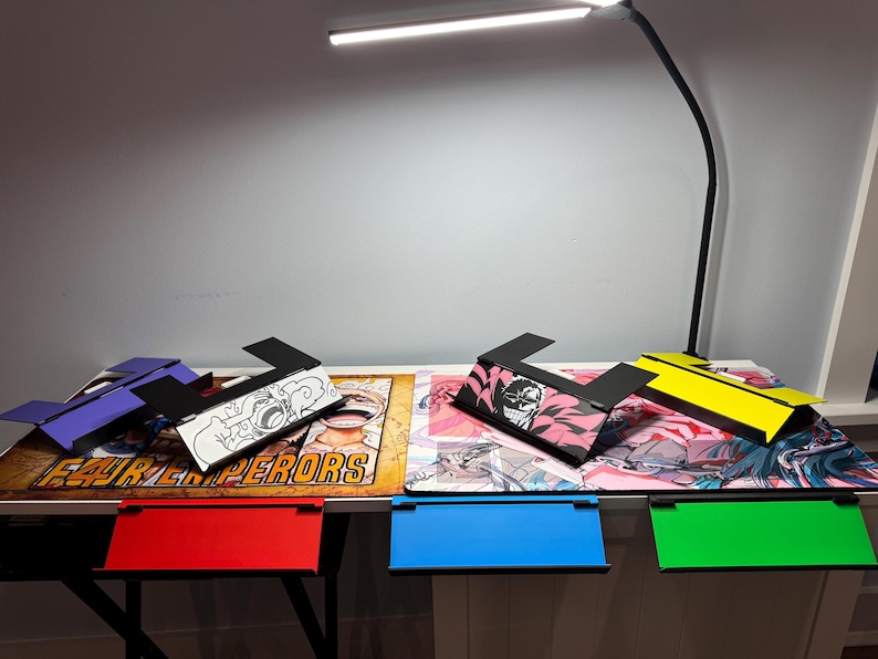 May include: Five different colored laptop stands with various anime designs. The stands are made of cardboard and fold flat for easy storage. The stands are on a table with a white surface. The text "FOUR EMPERORS" is visible on the table.