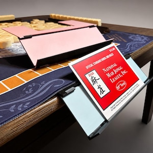 Puede incluir: Una mesa de Mahjong de madera con una superficie de juego azul y un estante rosa para fichas. La mesa presenta un folleto rojo y blanco con el texto "NATIONAL MAH JONGG LEAGUE, INC." y "2026". Se ven fichas de Mahjong beige.