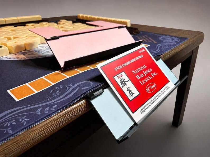 Puede incluir: Una mesa de Mahjong de madera con una superficie de juego azul y un estante rosa para fichas. La mesa presenta un folleto rojo y blanco con el texto "NATIONAL MAH JONGG LEAGUE, INC." y "2026". Se ven fichas de Mahjong beige.