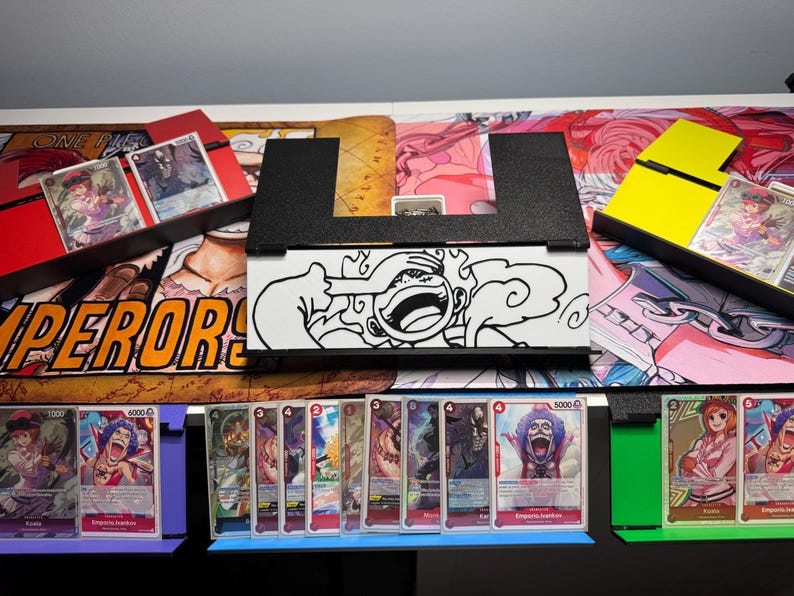 May include: A black card game deck box with a white cartoon image of a character with a cloud behind them. The box is sitting on a table with a colorful playing mat and several rows of trading cards. The cards are arranged in rows and have different images of characters from the One Piece anime series. The cards have the text "Emperor's" and "5000" printed on them.