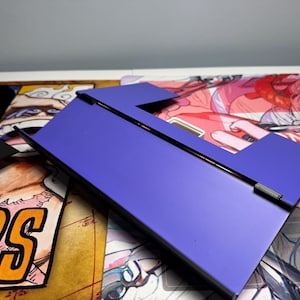 May include: A purple phone stand with a black hinge. The stand is made of a sturdy material and has a sleek design.