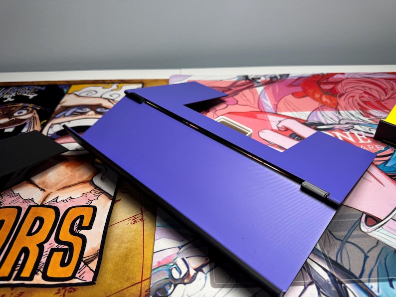 May include: A purple phone stand with a black hinge. The stand is made of a sturdy material and has a sleek design.