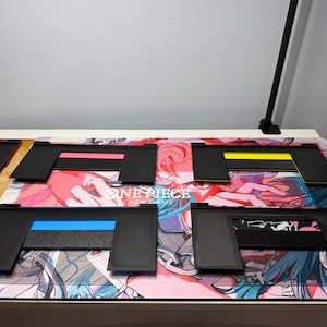 May include: Five black plastic card game holders with different coloured stripes. The holders are on a pink, blue and white anime-themed playmat with the text "ONE PIECE CARD GAME".
