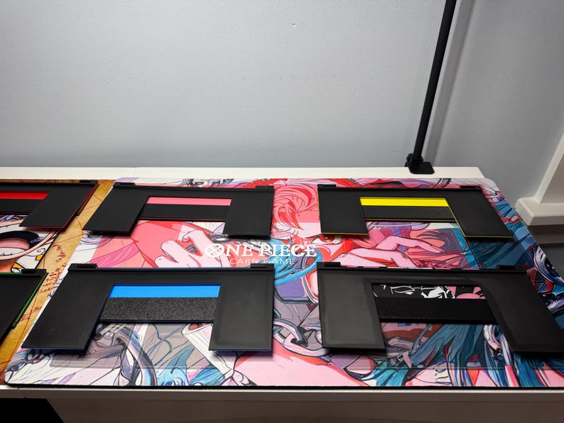 May include: Five black plastic card game holders with different coloured stripes. The holders are on a pink, blue and white anime-themed playmat with the text "ONE PIECE CARD GAME".