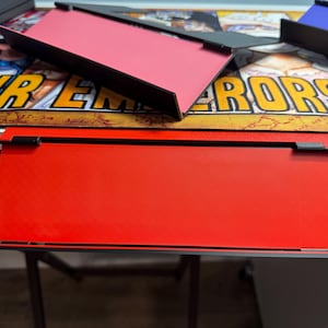 May include: A red, rectangular, flat surface with a black border. The surface is raised on two black supports. The surface is on a white table with a colourful background.