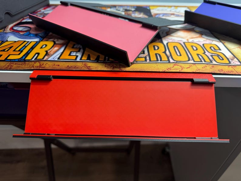 May include: A red, rectangular, flat surface with a black border. The surface is raised on two black supports. The surface is on a white table with a colourful background.