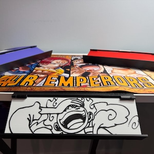 May include: A black and white illustration of a cartoon character with a large smile and a cloud behind them. The illustration is on a white background and is framed in a black frame. The frame is attached to a white desk with a black and orange anime-themed mousepad underneath.